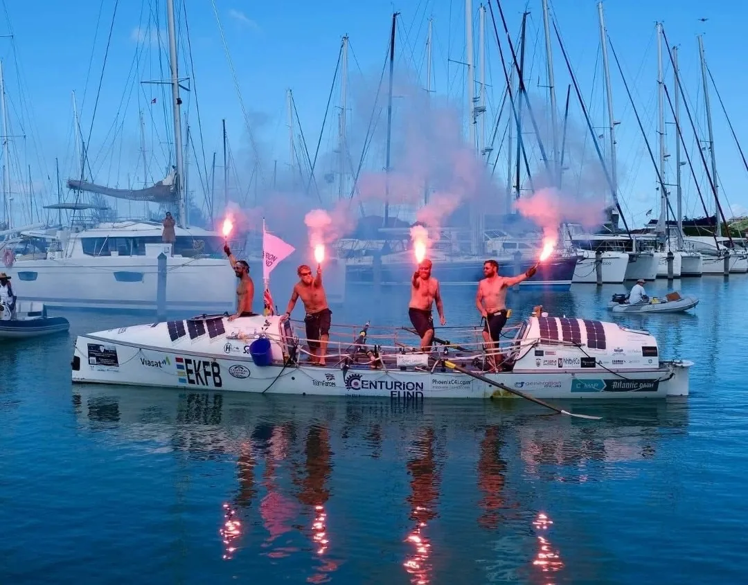 Four rowers holding pink flares aloft on their ocean rowing boat as they arrive in a marina in Antigua