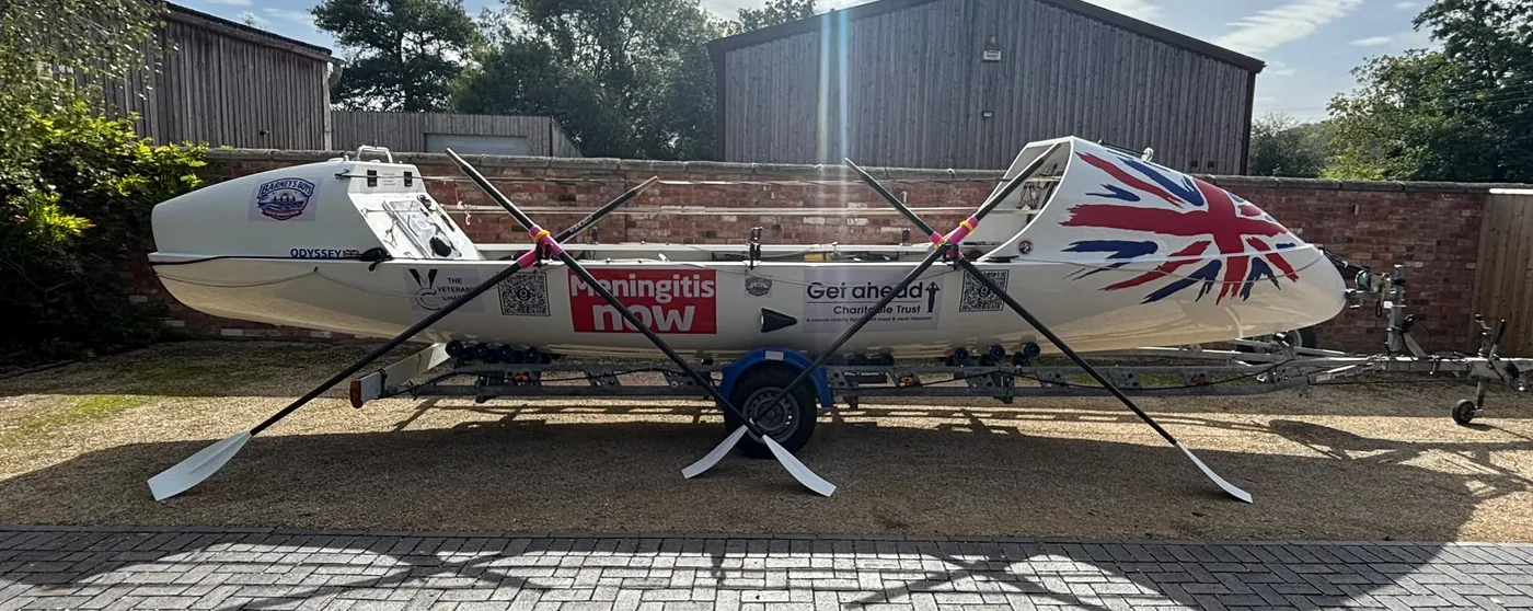 Odyssey on her trailer with two pairs of oars crossed in front, the Meningitis Now, Get A-Head and Veterans Charity logos on her hull